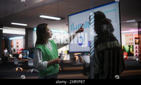 Two Creative Colleagues Collaborating, Pointing at Television Set With Financial Graphs And Charts in Office. Asian Female CEO Consults Black Marketing Manager. Two Women Brainstorming Stock Photo
