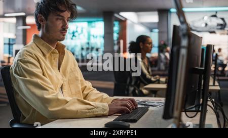 Male Caucasian Game Programmer Coding On Desktop Computer In Game Design Studio Diverse Office. Focused Man Writes Lines Of Code, Does Gameplay Engineering For New Immersive 3D Survival Video Game. Stock Photo