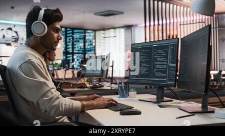 Male Indian Game Programmer Coding On Desktop Computer In Game Design Studio Diverse Office. Focused Man Writes Lines Of Code, Does Gameplay Engineering For New Immersive 3D Survival Video Game. Stock Photo
