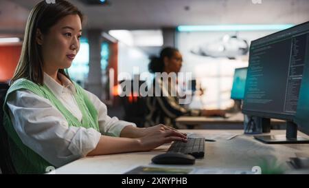 Female Asian Game Programmer Coding On Desktop Computer In Game Development Studio Diverse Office. Focused Woman Writes Lines Of Code, Does AI Engineering For New Immersive 3D Survival Video Game Stock Photo