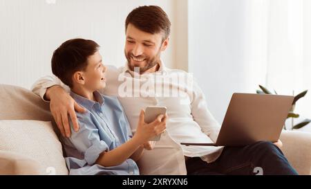 Cheerful Father And Son Using Laptop And Smartphone At Home Stock Photo