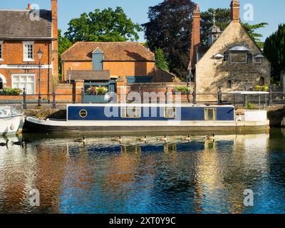 Saint Helen's Wharf is a noted beauty spot on the River Thames, just upstream of the medieval bridge at Abingdon-on-Thames. The wharf was for centuries an important transport and shipping link up the Thames and between canals from Oxford and the Midlands. Fine merchant houses rub shoulders with alms houses and the Saxon-era Saint Helens Church. Here we see pleasure and house boats moored alongside the Long Alley Alms Houses,  viewed on a fine mid-summer day from the north bank of the river. And a line of Canada Geese chooses this moment to swim past... Stock Photo