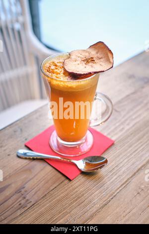 A warming glass of a drink topped with a sliced dried apple, served on a red napkin beside a silver spoon, set against a light wooden table and a blur Stock Photo