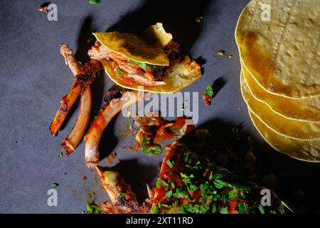 Top view of deconstructed rib tacos featuring spiced meat on skeleton ribs, garnished with fresh herbs and salsa. Corn tortillas stacked aside on a da Stock Photo