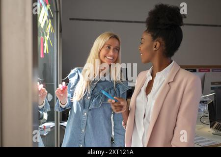 Smiling multiracial female colleagues standing near glass wall with sticky notes and working on project together while looking at each other Stock Photo