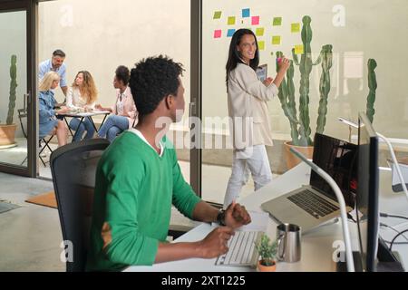 Black man using computer and cheerful woman writing on sticky notes during work on project in workspace Stock Photo