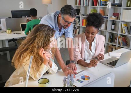 High angle of multiethnic coworkers gathering at table and speaking about new business strategy while discussing diagram on paper in workspace Stock Photo