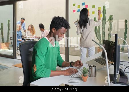 Side view of African American male office worker sitting at table and typing on keyboard while working in workplace with diverse colleagues Stock Photo