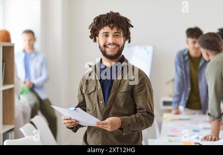Portrait of smiling mixed race man with dreadlocks standing with documents on workplace in office. Stock Photo