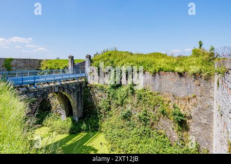 Footbridge at Charlemont fortress, ditch with stagnant water with blooming algae, brick walls covered by wild vegetation, sunny summer day with blue s Stock Photo