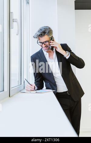 An elegant executive talking on his mobile phone, next to a window, symbolising connected businesses Stock Photo