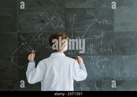 Back view of schoolboy in eyeglasses and white uniform standing while writing chemical formulas on chalkboard in classroom Stock Photo