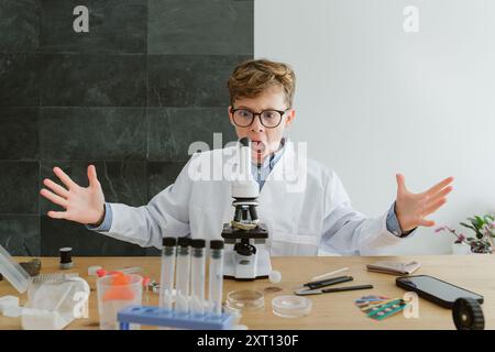 Schoolboy wearing glasses and lab coat sitting at table with various test tubes and surprised face expression while looking through microscope in labo Stock Photo