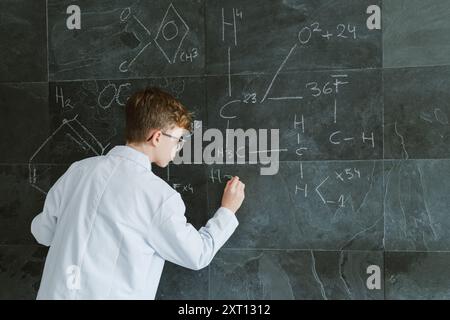 Back view of schoolboy in eyeglasses and white uniform standing while writing chemical formulas on chalkboard in classroom Stock Photo