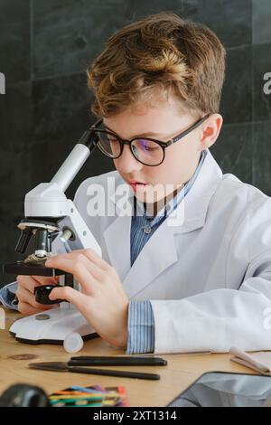 Preteen boy wearing uniform examining test tube with microscope while sitting at table and preparing for research in laboratory at school in classroom Stock Photo