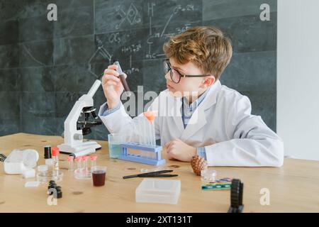 Focused preteen schoolboy in glasses and uniform conducting chemical experiment with test tube while sitting at desk near microscope against chalkboar Stock Photo