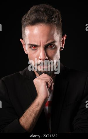 Intense young businessman in a black suit stares confidently with a hand on his chin against a dark background and red contact lenses Stock Photo