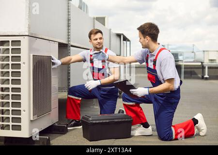 Team of young professional technicians sitting on rooftop and installing air conditioner. Stock Photo