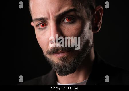 A close-up portrait of a bearded man with a stern expression wearing red contact lenses, illuminated by a dramatic side light that contrasts sharply w Stock Photo