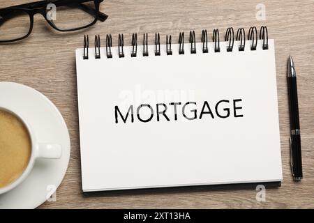 Word Mortgage written in notebook, eyeglasses, pen and coffee on wooden table, flat lay Stock Photo