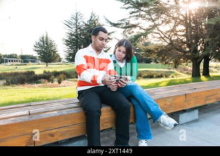 A young Latino couple sits closely on a park bench, engaged in viewing a tablet together during a sunny day, surrounded by greenery Stock Photo