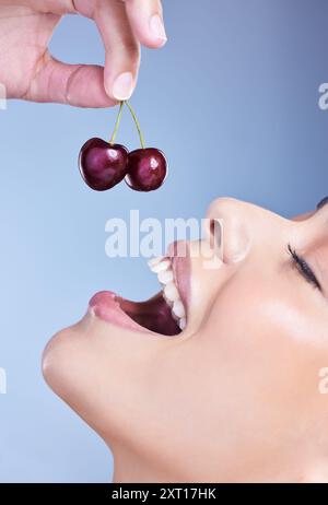 Woman, smile and eating cherry in studio with closeup for healthy nutrition, digestion and benefits. Mouth, happy and female person with fruit on blue Stock Photo