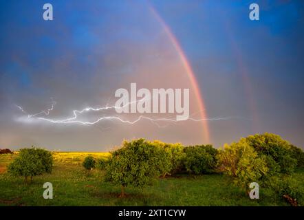 Vivid double rainbow arcs across a stormy sky while lightning strikes illuminate the horizon above a lush, green landscape. Dumas, Texas United States Stock Photo