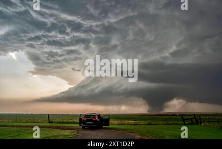 A storm chaser s vehicle parked on a dirt road observing a massive tornado forming under a dark, dramatic sky in an open field. Olustee, Oklahoma Unit Stock Photo