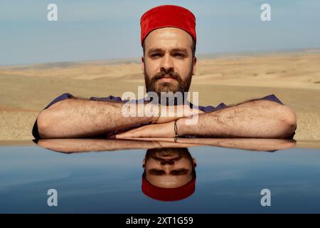 Man with a beard wearing a red fez hat rests his chin on his crossed arms, reflected on a glossy surface against a desert backdrop. bola02513 Copyrigh Stock Photo