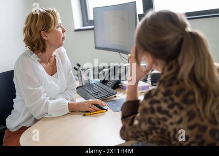 Diverse situatie in een huisartsenpraktijk Two women engaged in a conversation at an office desk with a computer monitor in the background. Molenhoek Stock Photo