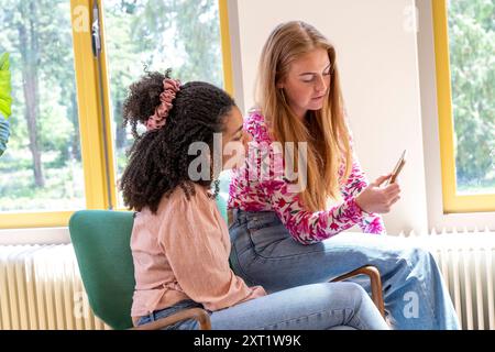 Two young women sitting indoors, one holding a smartphone and showing it to the other, both are engaged and seem to be having a friendly conversation. Stock Photo