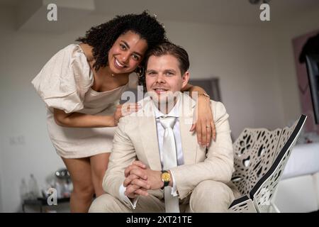 Smiling couple dressed in formal attire posing for a photo with a laptop in the foreground, indicating a festive or significant occasion at home. panc Stock Photo