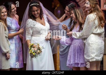 A radiant bride in a white lace dress smiles joyfully, surrounded by her bridesmaids in pastel attire, as they admire her gown and share a special mom Stock Photo