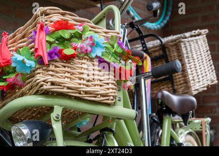 A close-up of a woven bike basket adorned with colorful flowers, mounted on a green bicycle, with another bicycle and brick wall in the background. pa Stock Photo