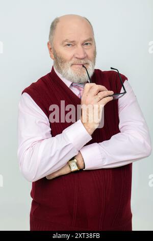 Portrait of an elderly gray-haired man with a beard and bald head. He is holding glasses and looking at the camera. Stock Photo