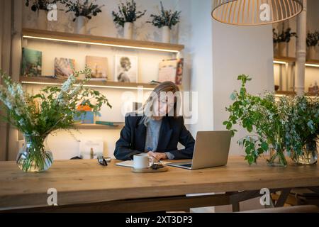 Businesswoman writing business plan, while working on laptop in office, thinking about strategies Stock Photo