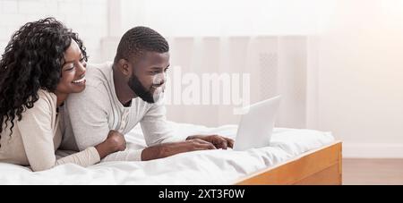 Cheerful afro spouses lying in bed using laptop together Stock Photo