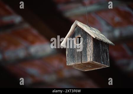 A charming weathered wooden birdhouse dangles from a string, set before the blurred warmth of a rustic red-brown roof The scene encapsulates the quain Stock Photo