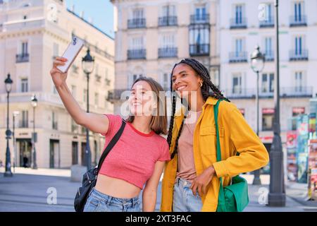 Two young multiethnic female friends take a cheerful selfie together on a bustling city street. Both women are looking at the smartphone, smiling broa Stock Photo