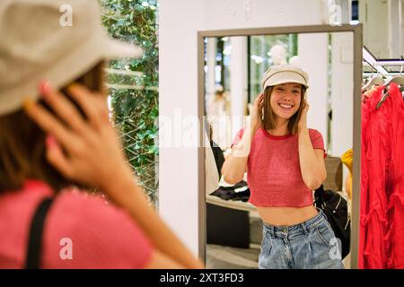 A cheerful young woman, smiling at her reflection in the mirror, tries on a stylish cap in a fashion boutique. She is dressed casually in a pink top a Stock Photo