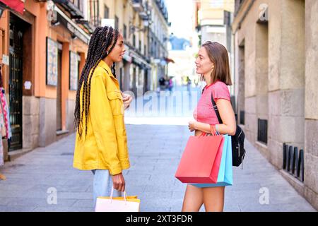 Side view of two young multiethnic women, each carrying shopping bags, engaged in a conversation looking at each other while walking down a bustling c Stock Photo