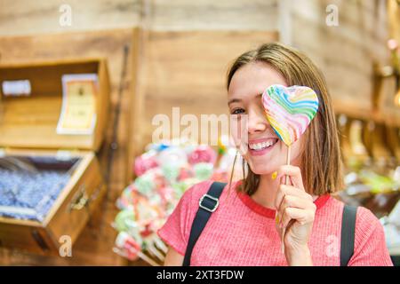 A happy young Caucasian woman is smiling at the camera, holding a large, heart-shaped, multicolored lollipop inside a vibrant candy store. Stock Photo