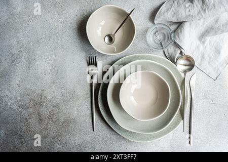A tastefully decorated minimalist table featuring a stack of muted gray plates, a small bowl, silver cutlery and a linen napkin on a textured gray bac Stock Photo