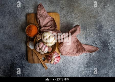Fresh whole garlic bulbs alongside spices on a wooden cutting board with a textured gray background A linen cloth adds a touch of warmth to the compos Stock Photo