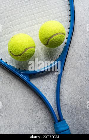 Close-up view capturing two bright yellow tennis balls beside the curve of a blue racket handle, all set against a textured gray background Stock Photo