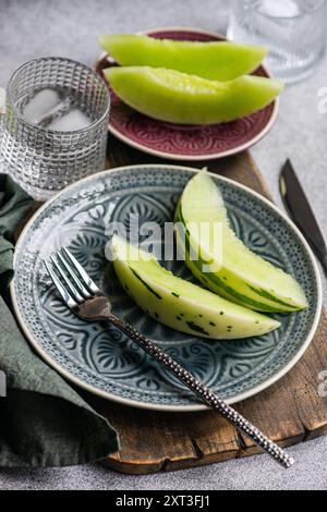 From above organic mini melon slices arranged on ornate ceramic plates, accompanied by a glass of water with ice and a fork on a textured tablecloth. Stock Photo