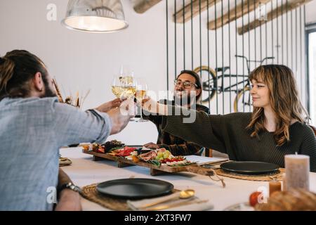 In a warmly lit dining room, three friends, two men and a woman, enjoy a delightful dinner prepared by a private chef. The friends are toasting with g Stock Photo