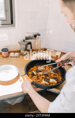 From above a private chef, visible from the shoulders down, expertly prepares a succulent fish dish in a skillet. He works in a well-lit, cozy kitchen Stock Photo