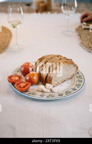 Elegantly laid dining table featuring freshly sliced bread on a decorative plate, complemented by ripe tomatoes and garlic cloves, with wine glasses i Stock Photo