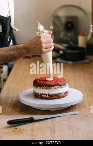 Cropped unrecognizable male chef expertly applies cream to a layered red velvet cake in a bright kitchen, focus on the hands wielding a piping bag. Stock Photo